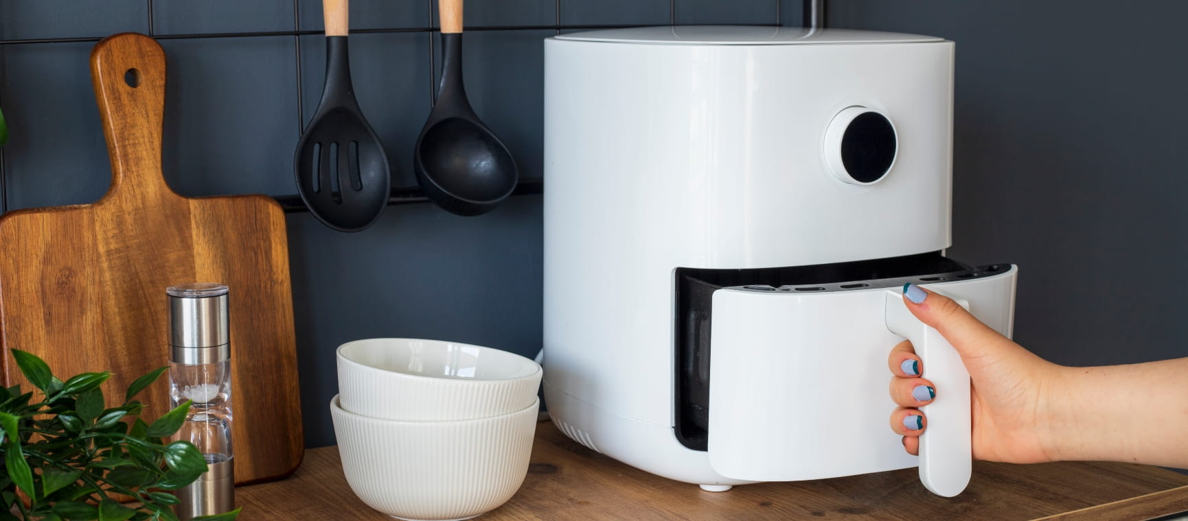 A hand opening the drawer of a white air fryer which is on a wooden kitchen counter against a navy blue wall, with other kitchen tools visible