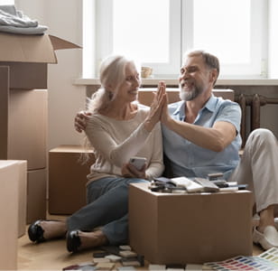 Two older people sitting on the floor surrounded by boxes, high fiving